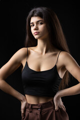 Classic studio portrait of a young brunette dressed in a black top, who is sitting on a chair against a black background.