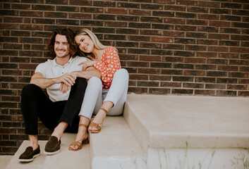 Smiling young couple in love sitting in front of house brick wall