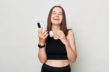 Wall Mural - Laughing happy cheerful woman wearing black top standing isolated over white background holding cell phone looking at camera reading messages in social networks.