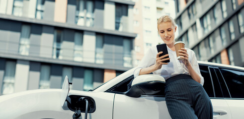 Focus businessman using phone, leaning on electric vehicle, holding coffee with blurred city residential condo buildings in background as progressive lifestyle by renewable and sustainable EV car.