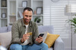 © Tetiana - Smiling young hispanic man sitting on sofa at home in hotel and using credit card and phone