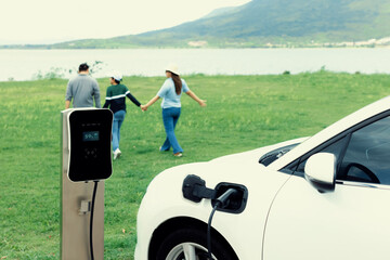 Concept of progressive happy family enjoying their time at green field and lake with electric vehicle. Electric vehicle driven by clean renewable from eco-friendly power sauce.