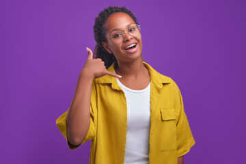 Wall Mural - Young positive attractive African American millennial woman making call me gesture and smiling looking at camera inviting to talk on mobile phone after work stands on plain purple background.