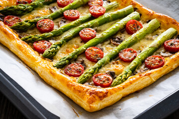 Poster - Baked puff pastry with green asparagus with mozzarella cheese, mushrooms and cherry tomatoes on baking paper on wooden table
