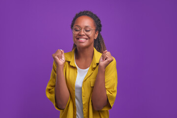 Young happy curly African American woman in glasses closing eyes and making victory gesture with both hands rejoicing at fulfillment of cherished dream or achievement of goal stands in purple studio.