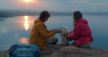 Cheerful Couple With White Spitz Dog Sit On Top Of Mountain Enjoying Time During Travel, Pretty Caucasian Family In Sportswear Has Fun With Pet At Sunset In The Evening. Tourism, Travel, Adventure