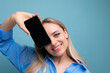 smiling stylish blond girl holding smartphone screen with mockup for web page near her face on blue background