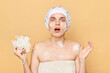 Shocked astonished surprised female dressed bath cap looking at camera with open mouth holding sponge with foam taking shower in thew morning expressing fear.