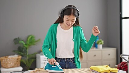 Canvas Print - Young chinese woman listening to music ironing clothes dancing at laundry room