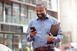 © Matthew C/peopleimages.com - Businessman, phone and typing an email at office, building and corporate workplace communication. Happy, senior and black man on mobile, smartphone or technology for online search, text or contact