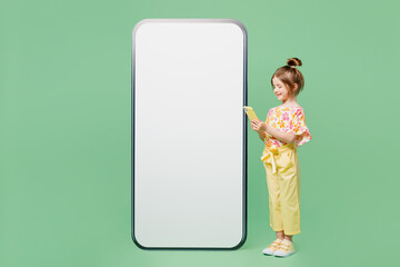 Wall Mural - Full body side view little child kid girl 6-7 years old wear casual clothes big blank screen area mobile cell phone use smartphone isolated on plain green background. Mother's Day love family concept.