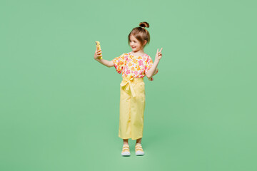 Wall Mural - Full body little child kid girl 6-7 years old wear casual clothes doing selfie shot on mobile cell phone e show v-sign isolated on plain green background. Mother's Day love family lifestyle concept.
