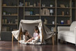 Happy mom and adorable little child girl having fun in play tent, resting on heating floor in handmade toy fort from chairs and blanket, talking, chatting, laughing
