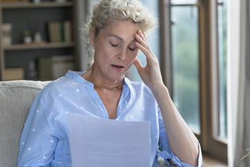 Shocked senior elder woman holding paper legal document, sitting on home sofa, touching head with closed eyes, feeling stress, despair, thinking on bad health problems, bankruptcy, financial crisis