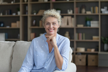 Wall Mural - Positive beautiful mature blonde woman posing for shooting on couch in apartment, touching chin, looking at camera with toothy smile. Senior homeowner lady home head shot portrait