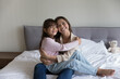 © fizkes - Cheerful pretty mom holding cute preschool kid girl in arms, hugging child, sitting on bed, looking at camera, smiling, enjoying home comfort, motherhood. Mother and little daughter portrait