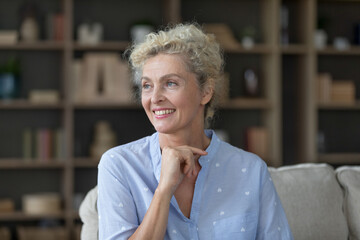 Wall Mural - Cheerful pretty mature woman relaxing at home, sitting on sofa, looking at window away with toothy smile, dreaming thinking on good future plans, touching chin. Positive senior lady casual portrait