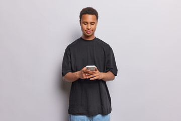 Wall Mural - Horizontal shot of dark skinned man types text messages via smartphone uses contemporary technology dressed in casual black t shirt and jeans isolated over white background. Online communication