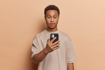 Sticker - Surprised dark skinned young man looks shocked at mobile phone reads message with amazed face expression receives stunning news dressed in casual t shirt isolated over brown background says wow