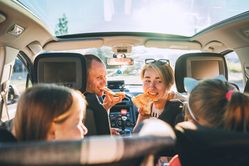 Young couple with daughters eating just cooked Italian pizza sitting in modern car with transparent roof. Happy family moments, childhood, fast food eating or auto journey lunch break concept image.