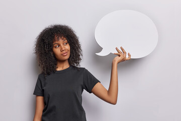 Pensive dreamy curly haired woman looks mysteriously at communication bubble thinks about advertising content wears casual black t shirt isolated over white background. Place your idea or text here