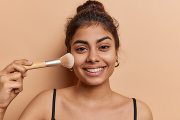 Pretty young dark haired woman radiates joy as she smiles at camera while applying makeup with brush to create stunning look wears t shirt has healthy clean skin isolated over brown background.