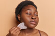 © WHstudio Leushin N - Plump woman with perfect glowing skin massaging face by guasha stone scraper improves circulation keeps eyes closed stands bare shouldered isolated over brown background. Female beauty trends