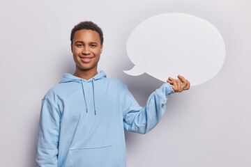 Wall Mural - Pleased delighted young African man holds empty speech bubble with mock up space for text dressed in casual blue hoodie suggests to place your advertisement here stands against white background.