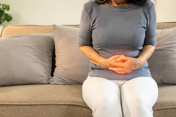 Asian old woman sitting on sofa with stomach ache irritable bowel syndrome Abdominal pain in the elderly, flatulence, inflammatory bowel disease, health care