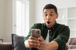 Happy excited African American man holding a mobile phone in his hands, looking at the screen with a surprised face, young male reading good news sitting at home on the couch
