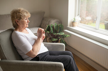 senior female in her 70s holding smartphone using mobile app, texting message, search ecommerce offers on cell phone technology device sitting in a chair at home.