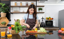 Young Asian Housewife Dressed In An Apron Slice Bell Pepper, Tomato And Crisphead Lettuce On A Wooden Chop Board. The Kitchen Counter Full Of Various Kinds Of Fruits And Vegetables.