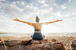 © Westend61 - Woman practicing yoga with arms outstretched sitting on rock at beach in front of sky