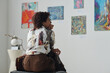 © pressmaster - Rear view of African American woman sitting on couch and looking at paintings on the wall in gallery