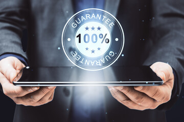 Poster - Close up of businessman hand holding pad with glowing 100% guarantee stamp hologram on dark background with bokeh circles. Quality seal and warranty concept.