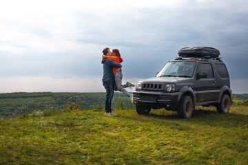 Happy couple kissing and hugging standing on the top of the mountain. Car trip on SUV with roof rack, trip at summer day. Man and woman enjoying time together travelling by vehicle. Copy space