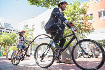 Businessman father in suit riding tandem bicycle son on urban sidewalk