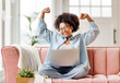© JenkoAtaman - beautiful young smiling ethnic woman smiling and rejoices in victory   sitting on sofa and working at laptop screen after finishing project  in home office