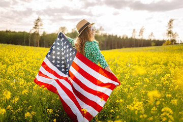 Wall Mural - Young woman with american flag on blooming meadow. 4th of July. Independence Day. Patriotic holiday. USA flag fluttering in the wind.