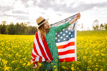 Wall Mural - Young woman with american flag on blooming meadow. 4th of July. Independence Day. Patriotic holiday. USA flag fluttering in the wind.