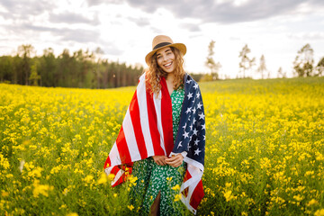 Wall Mural - Young woman with american flag on blooming meadow. 4th of July. Independence Day. Patriotic holiday. USA flag fluttering in the wind.