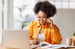 © JenkoAtaman - Beautiful young smiling ethnic woman making call via smartphone while working remotely from home while sitting at desk and talking to coworkers .
