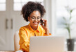 Beautiful young smiling ethnic woman making call via smartphone while working remotely from home while sitting at desk and talking to coworkers .