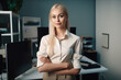 © ADDICTIVE STOCK - Generative AI illustration of smiling blond female manager in elegant shirt with crossed arms looking at camera while standing against office room