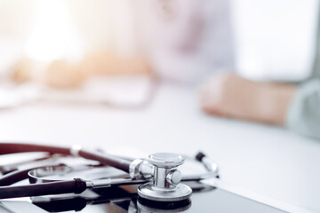 Doctor and patient are sitting and discussing something at the desk in the clinic office. The focus is on the stethoscope lying on the table, close up. Perfect medical service and medicine concept.