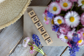 Daisy flowers in white cup composition with a straw hat on wooden background. Summertime, vacations, happiness concept. Flat lay, top view. Letters with word summer on a old book