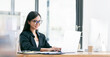 © NAMPIX - Portrait of young asian businesswoman with glasses smiling and working on laptop while sitting at office desk in modern office.