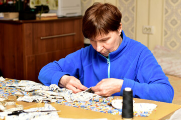 A seamstress sits at a table and sews two garments together with a needle and thread.