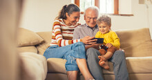 Happy Small Family Gathered Together, Sitting On A Sofa In The Living Room And Using A Smartphone. Mother, Son And Grandfather Watching Videos On A Mobile Phone And Laughing. They Are Bonding 