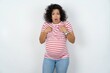 Embarrassed young pregnant woman wearing striped t-shirt over white indicates at herself with puzzled expression, being shocked to be chosen to participate in competition, hesitates about something
